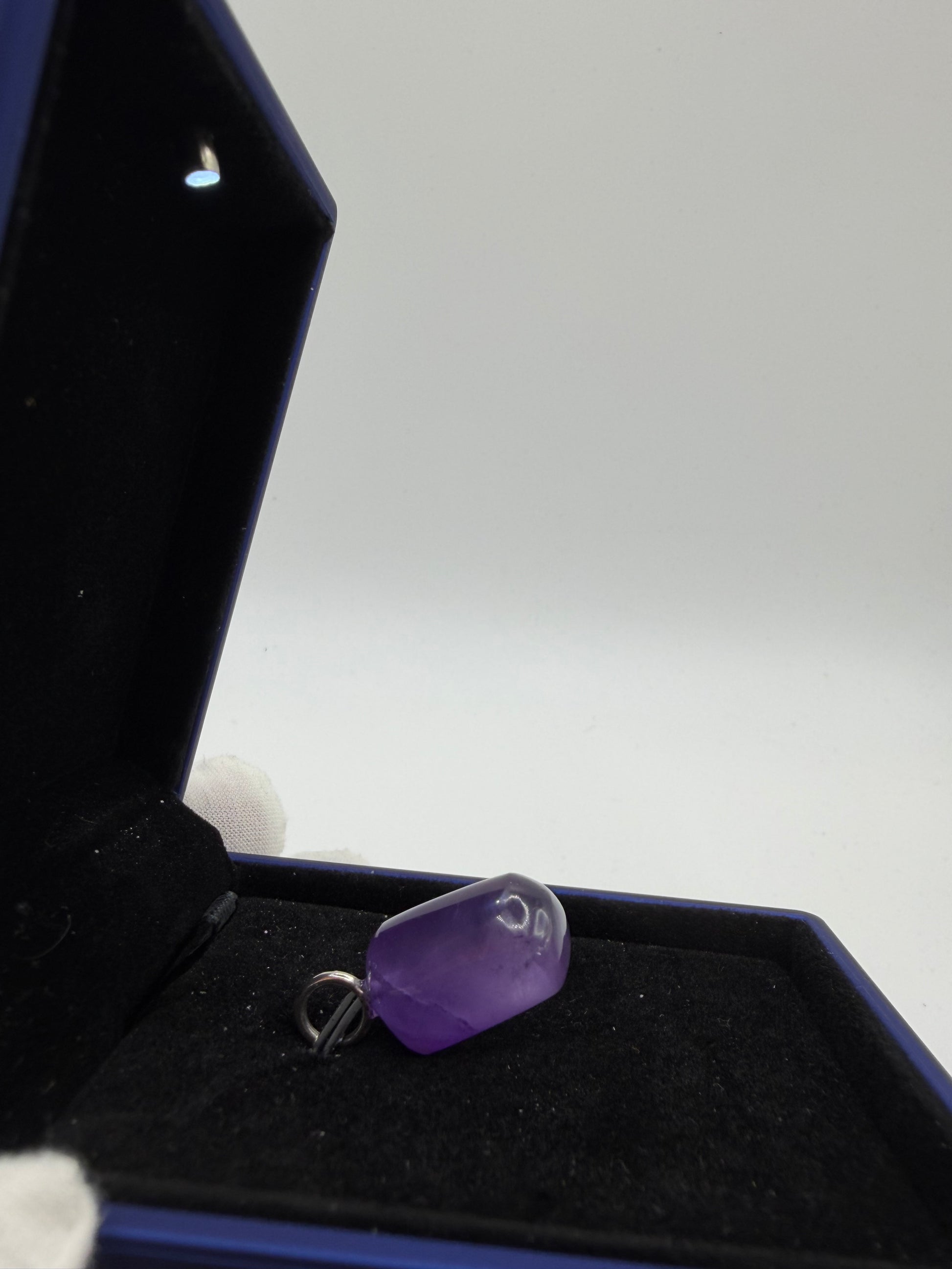 A polished purple gemstone pendant with a small silver metal loop, displayed inside an open dark blue jewelry box with black velvet lining.