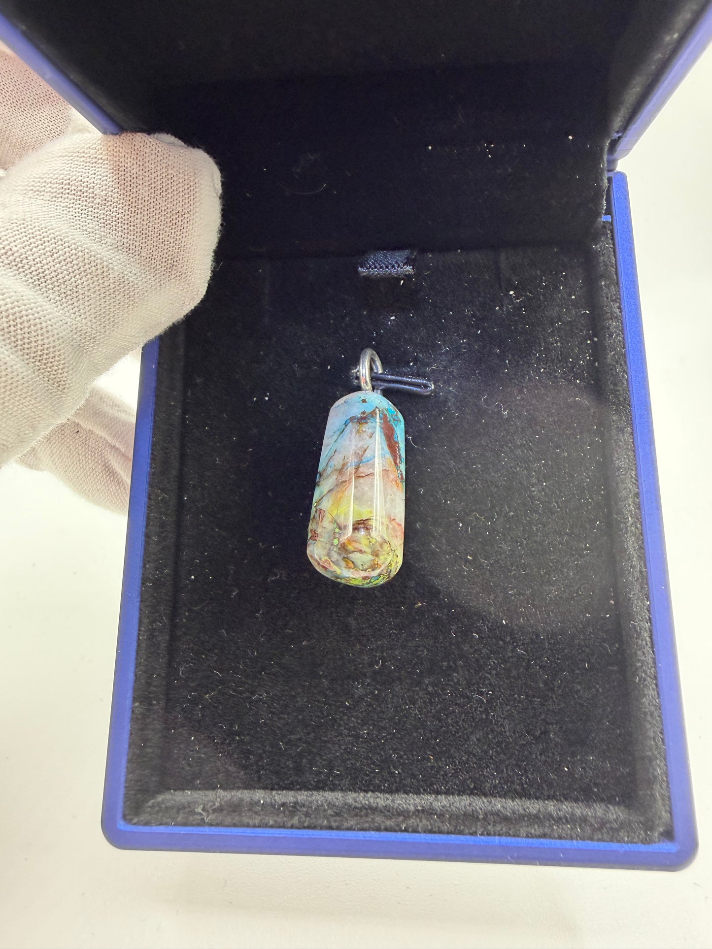 A small cylindrical pendant with a colorful marbled pattern in blue, yellow, and white, resting inside an open blue presentation box with a black velvet lining. A gloved hand is visible holding the box.