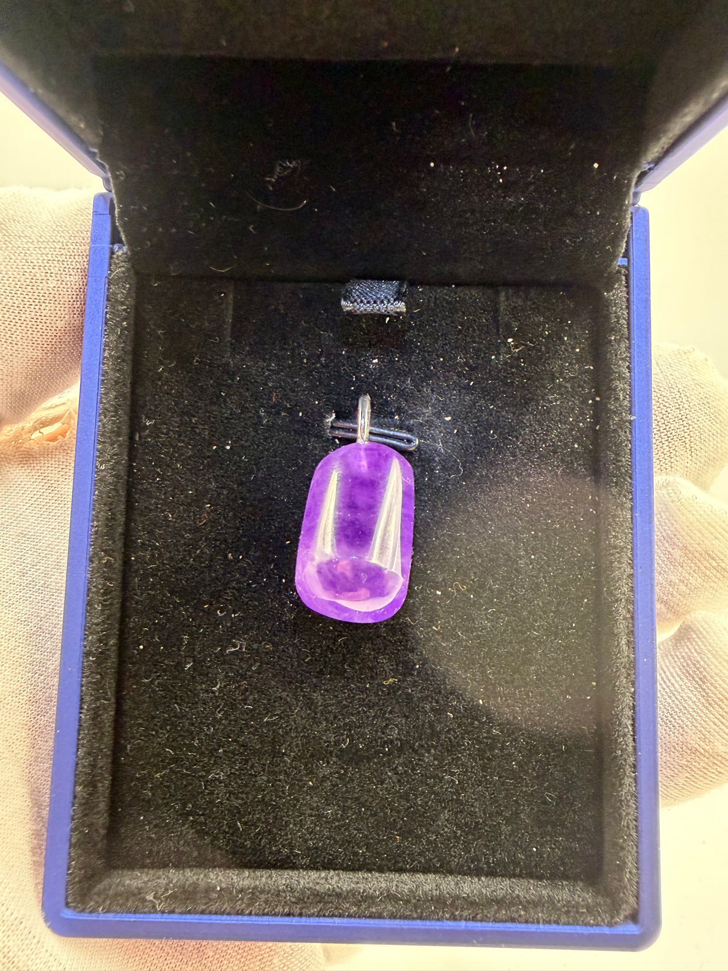 A small purple translucent capsule-shaped pendant or charm with a silver metal loop, displayed inside an open blue jewelry box with a black velvet interior.