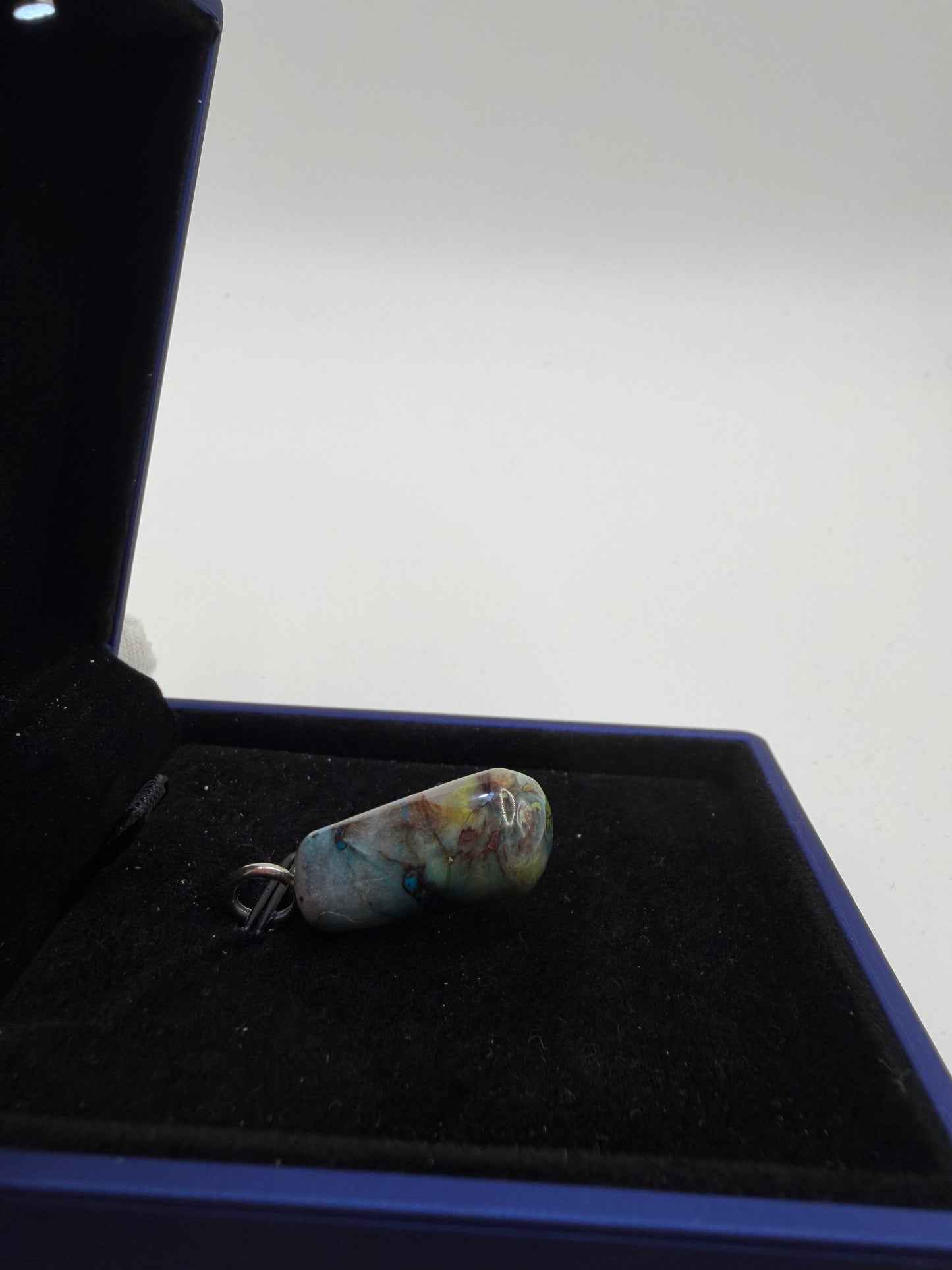 A small marbled pendant or charm with blue, green, and yellow tones, attached to a silver ring, displayed inside a black velvet-lined jewelry box.