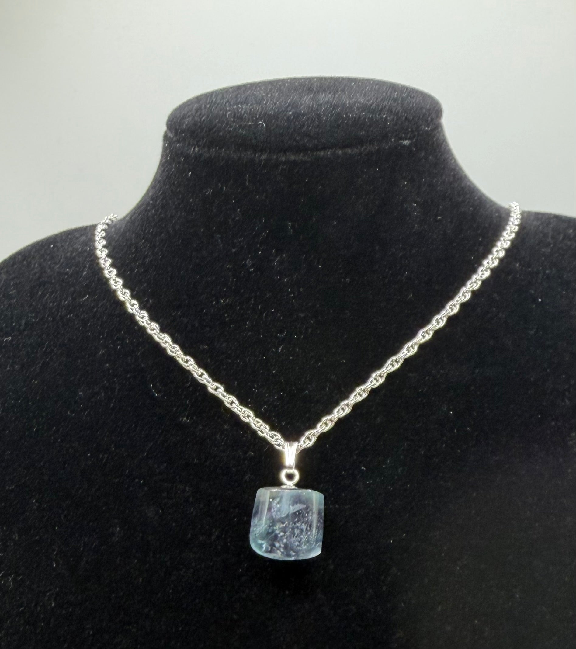 21ct Namibian fluorite pendant on black jewelry bust with sterling silver rope chain, showing icy aqua-blue cube-cut stone