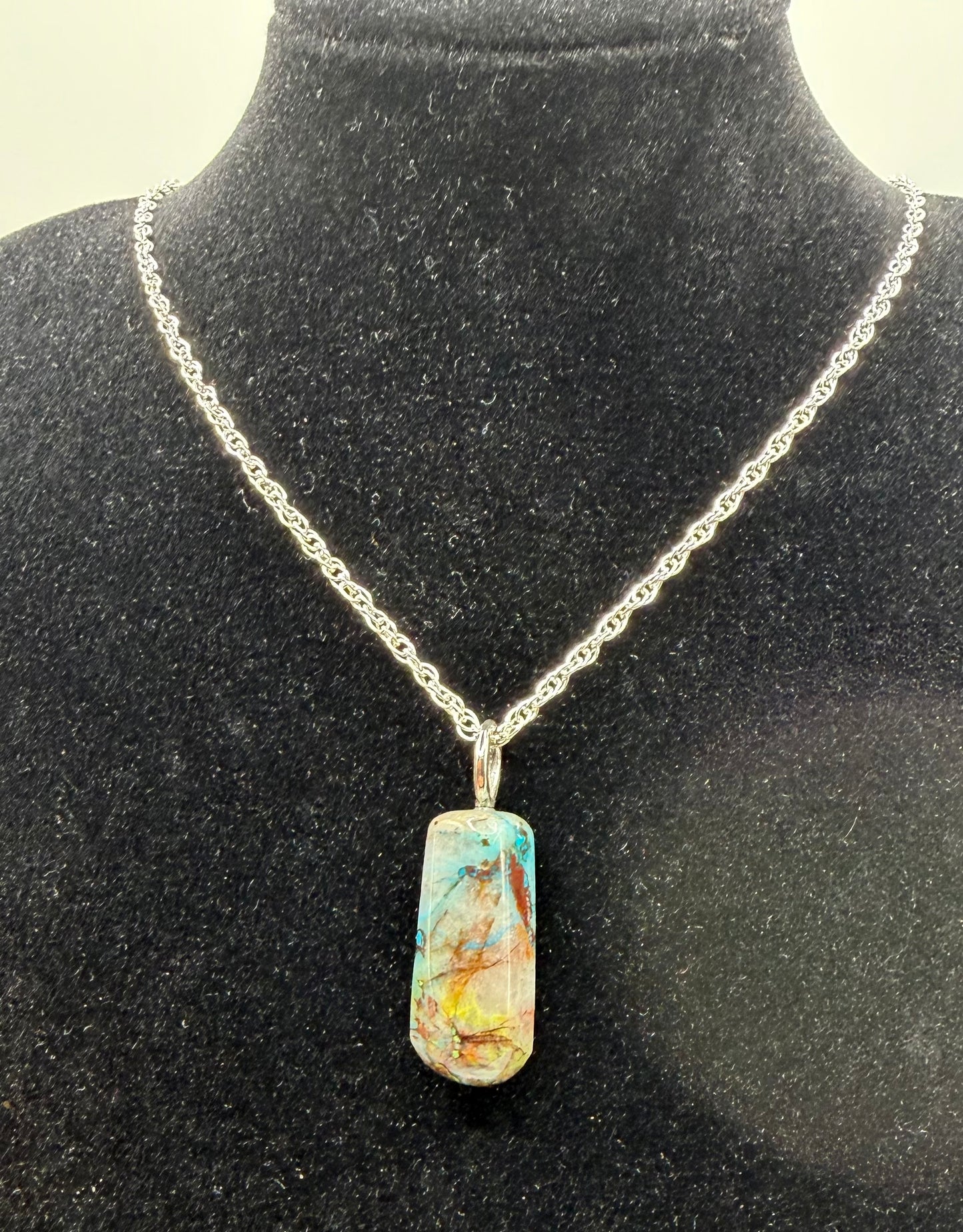 A silver chain necklace with a rectangular pendant featuring a marbled turquoise, gold, and brown pattern, displayed on a black velvet jewelry bust.