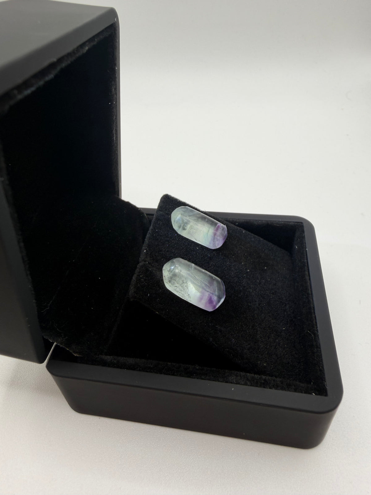 Natural Namibian fluorite stud earrings with near-colorless to violet-purple zoning in open black jewelry box, angled view