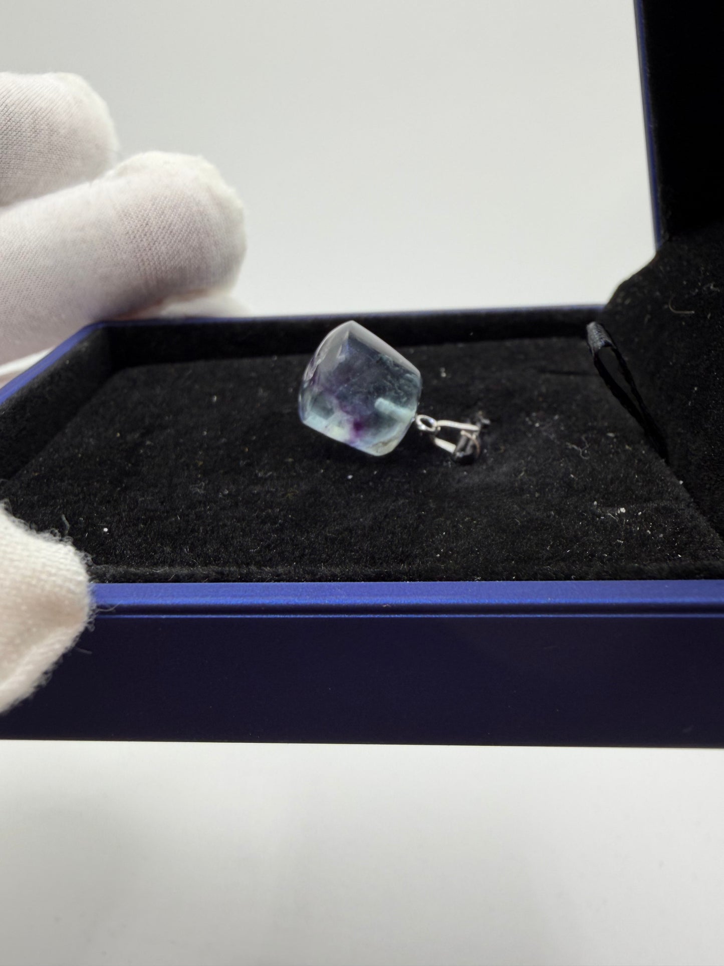 Gloved hand holding 21ct Namibian fluorite cube pendant over blue jewelry box, showing stone clarity and sterling silver peg bail