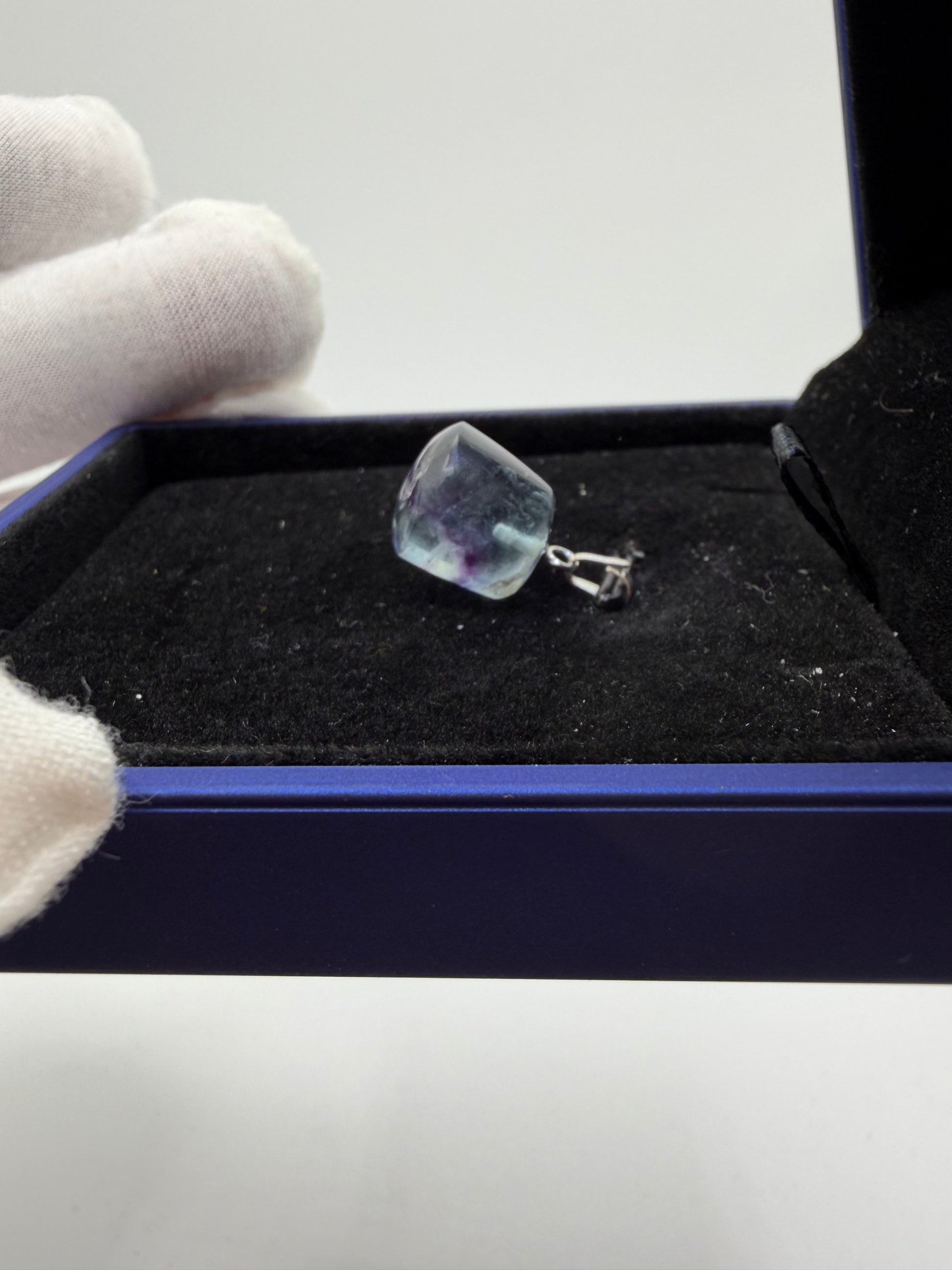 Gloved hand holding 21ct Namibian fluorite cube pendant over blue jewelry box, showing stone clarity and sterling silver peg bail