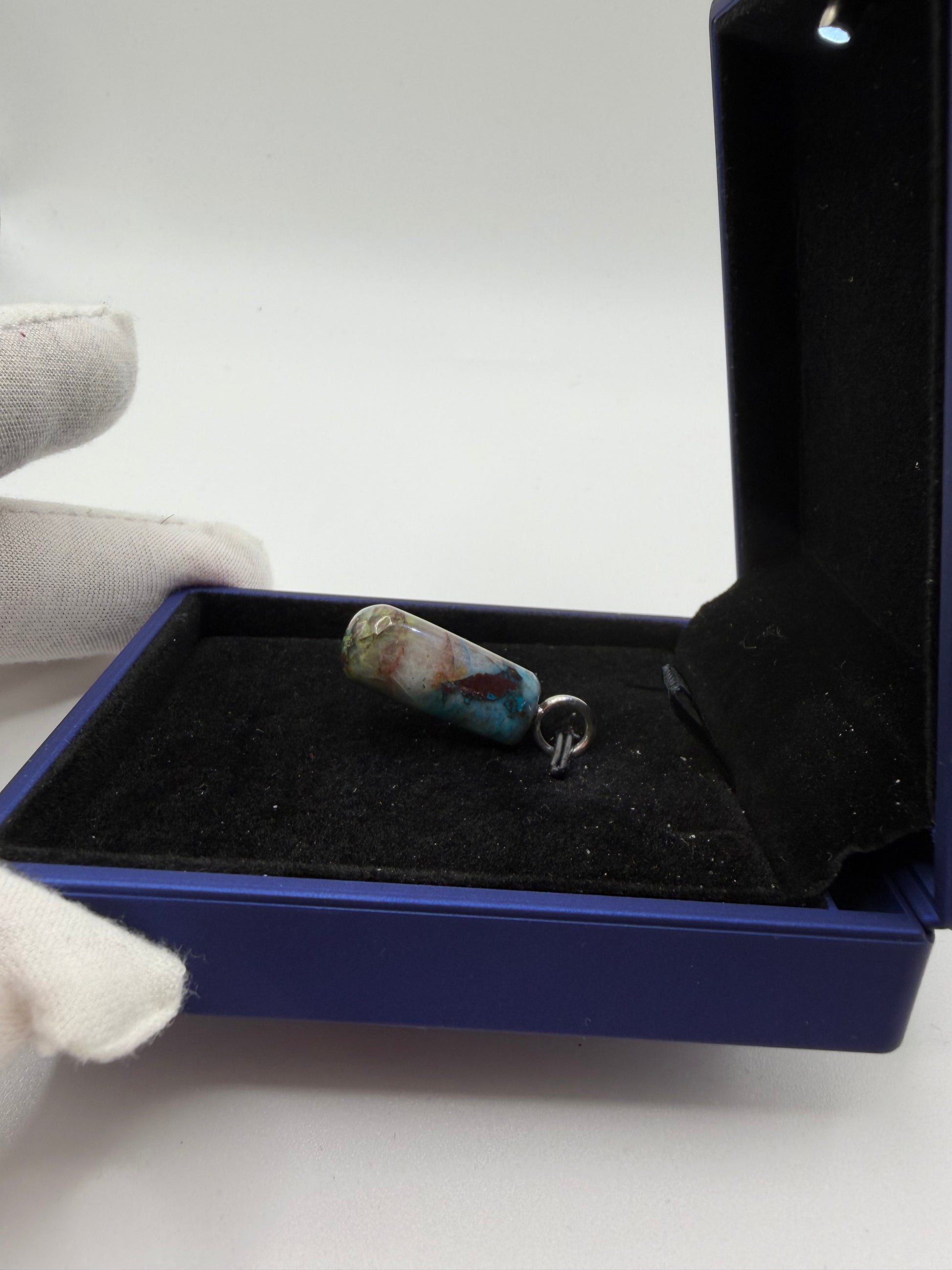 A small pendant with a turquoise-blue and cream marbled pattern on a silver bail, displayed inside an open dark blue jewelry box held by a white-gloved hand.